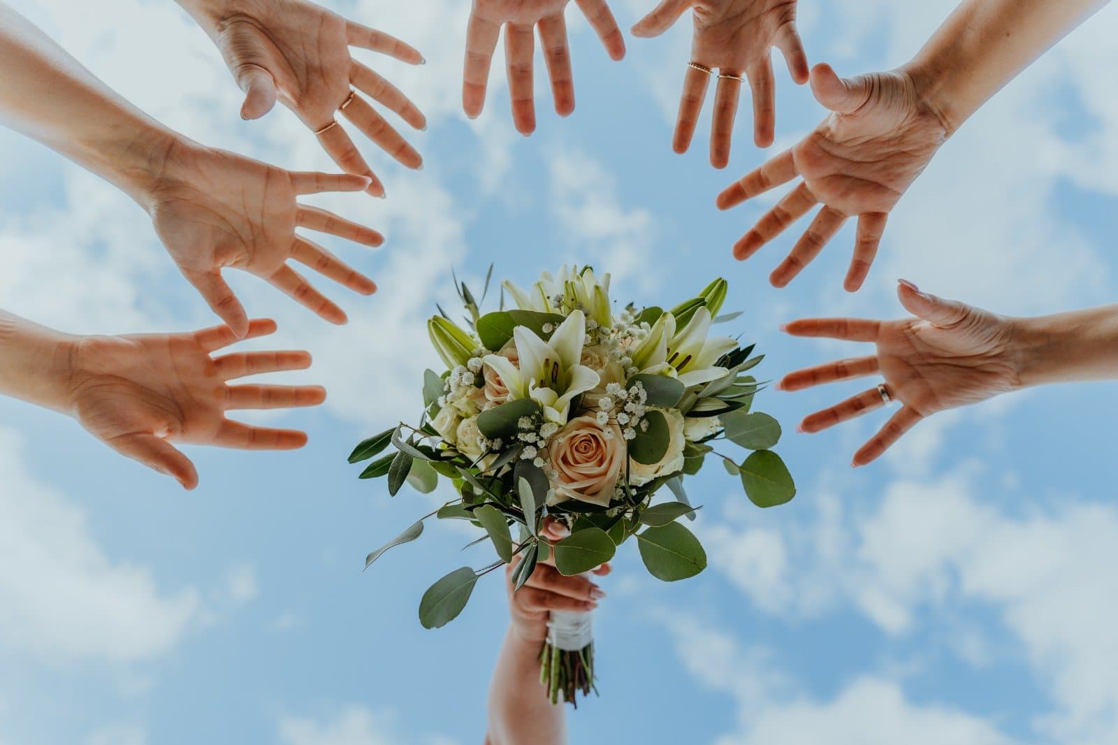 Mãos de amigas estendidas para o bouquet de lírios brancos, rosas cor de pêssego e eucalipto, erguido contra o céu